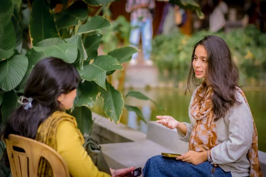 Two women sitting near lush plants having a conversation outdoors. Relaxed and candid atmosphere.